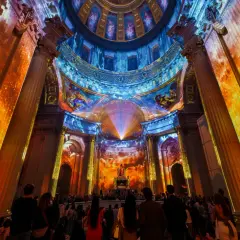 Spectacle lumineux projeté sur des colonnes et un dôme, créant une aura de couleurs vives. Une foule admire cette installation, comme si les dates invalides s'effaçaient.