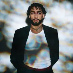 Sofiane Pamart in Berlin, mit dunklem Haar und Bart, trägt einen schwarzen Blazer über einem weißen Shirt. Bunte Lichtreflexe auf seinem Shirt.