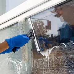 Osoba w niebieskich rękawiczkach myje okno gąbką i ściągaczką, tworząc pianę.