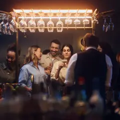 Glada gäster minglar och skrattar vid baren på Gothia Towers Skybar. Glas hänger i taket, redo för drinkar.