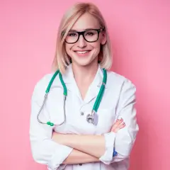 Uśmiechnięta pielęgniarka w okularach i białym fartuchu, z zielonym stetoskopem na szyi, stoi ze skrzyżowanymi rękami.