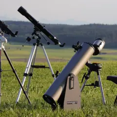 Trzy teleskopy na statywach stoją na trawiastym polu, w tle las i góry.