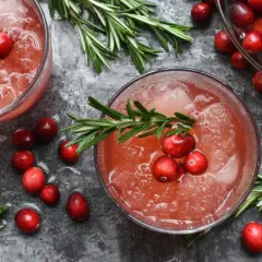 Dwa drinki z żurawiną i rozmarynem na ciemnym tle, otoczone świeżymi owocami i gałązkami.
