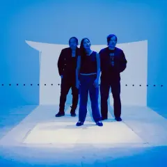 Trzy osoby stoją na scenie oświetlonej na niebiesko, tworząc atmosferę idealną do shoegaze.
