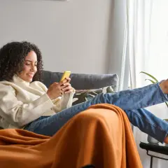 Frau mit lockigen Haaren entspannt sich richtig sitzend auf dem Sofa, schaut auf ihr Handy.