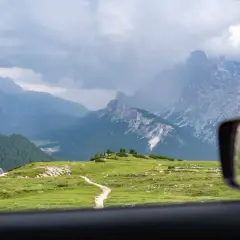 Vue depuis une voiture en Italie, traversant des prairies verdoyantes et des montagnes majestueuses sous un ciel nuageux.