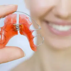 Dłoń trzyma pomarańczowy, wieloczynnościowy aparat dentystyczny. W tle uśmiechnięta twarz.