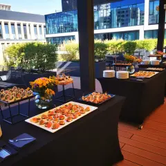 Elegancki catering Warszawa na imprezę plenerową. Stoły z przekąskami i napojami na tarasie z widokiem na nowoczesne budynki.