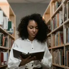Młoda kobieta z burzą loków czyta książkę w bibliotece. Szuka inspiracji, co napisać we wniosku o stypendium szkolne.