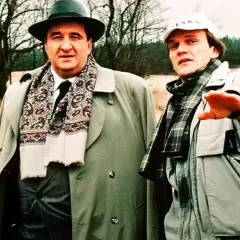 Dwóch mężczyzn w płaszczach i szalikach, jeden w kapeluszu, drugi w czapce. W tle drzewa i piasek. Nic śmiesznego.