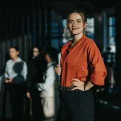 Uśmiechnięta kobieta w pomarańczowej bluzce trzyma teczkę, jakby właśnie odebrała prawo jazdy. W tle grupa osób.