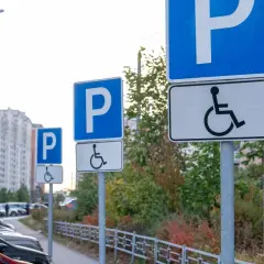 Znaki informują, komu przysługuje karta parkingowa. Na parkingu stoją samochody, a w tle widać budynki mieszkalne.
