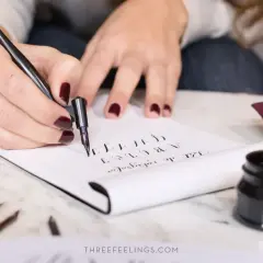 Manos practicando lettering con pluma y tinta, creando letras cursivas en un cuaderno.