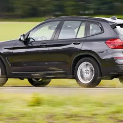 Ciemnoszary BMW G01 pędzi po wiejskiej drodze, rozmazując zielone pola i drzewa w tle.