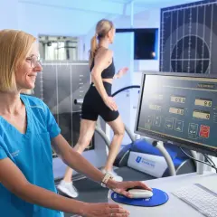 Frau in blauer Kleidung bedient Computer für Laufanalyse, während eine Läuferin auf Laufband trainiert.