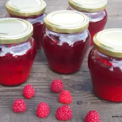 Słoiki z domowym sokiem z malin z sokownika, pełne lata. Kilka świeżych malinek leży obok.