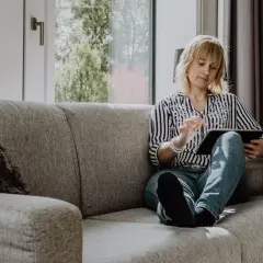Femme blonde sur un canapé gris, consultant une tablette. Elle semble chercher un avis sage-femme, peut-être pour une grossesse.