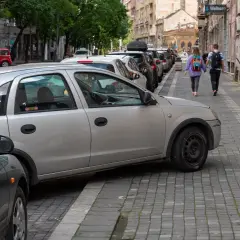 Samochody na chodniku utrudniają przejście. Dwie osoby idą wąskim chodnikiem, omijając przeszkody.