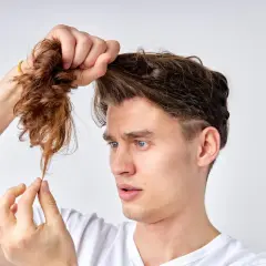 Mann mit blauen Augen betrachtet besorgt seine extrem kaputten Haare, die er in der Hand hält.