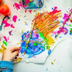 Kinderhand malt mit bunten Farben auf Papier, umgeben von Farbtöpfen und Pinseln.