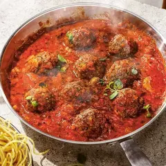 Frikadeller i tomatsås som puttrar i en panna, med en skål spaghetti bredvid.