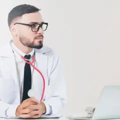 Lekarz w białym kitlu z czerwonym stetoskopem, gotowy do badań medycyny pracy. Obok laptop i lampa.