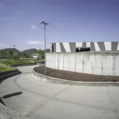Ampia rampa in cemento fai da te, curva dolcemente verso un edificio moderno con facciata a strisce bianche e grigie, circondata da verde e colline.