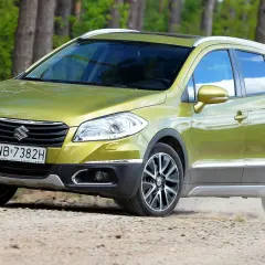 Zielony Suzuki S-Cross na drodze leśnej, wzbudzający pozytywne suzuki s-cross opinie. Kurz spod kół świadczy o jego terenowych możliwościach.