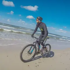 Rowerzysta przemierza piaszczyste plaże Słowińskiego Parku Narodowego. W tle błękitne niebo i fale Bałtyku.
