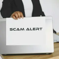 Ostrzeżenie przed oszustwami na Vinted. Ktoś trzyma kartkę z napisem "SCAM ALERT" nad laptopem.