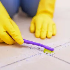 Hände in gelben Handschuhen reinigen mit einer lila Zahnbürste Fliesen und Fugen.