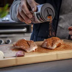 Ein Mann bestreut Hähnchenbrust mit einem magischen Gewürz-Rub auf einem Holzbrett. Zwiebeln und Knoblauch liegen daneben.