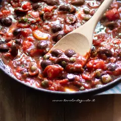 Un cucchiaio di legno mescola un sugo ricco e saporito, pieno di pomodori, olive nere e capperi. È la base perfetta per una pasta alla puttanesca.
