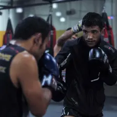 Two men in boxing gloves engage in a spar fight in a gym, with punching bags in the background.