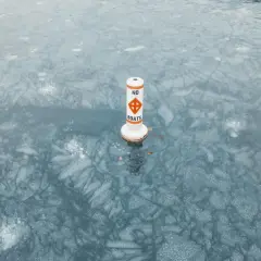 Bojka z napisem "NO BOATS" stoi na zamarzniętej tafli wody, ostrzegając przed ruchem łodzi.