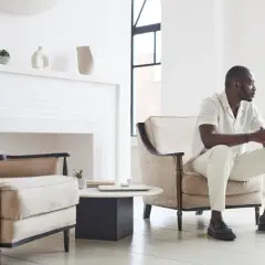 Un homme élégant en tenue claire, assis dans un fauteuil, montre comment porter des mocassins avec style.