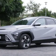 Nowy Hyundai Kona Hybrid w kolorze jasnoszarym, zaparkowany na brukowanym placu, otoczony zielenią.