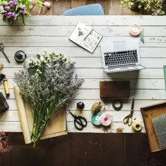 Kwiaty, laptop i akcesoria florystyczne, jak nożyczki, wstążki i sznurek, tworzą inspirujący warsztat. Widać też materiały do pakowania, przypominające **faszynę**.