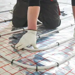 Ręce w białych rękawiczkach układają białe rurki na folii z siatką. To początek prac nad tym, jak się robi ogrzewanie podłogowe.