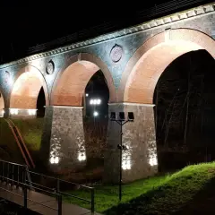 Nocna iluminacja kamiennego wiaduktu z ceglanymi łukami. Widać fragment mostku i drzewa. To piękna bytów trasa.
