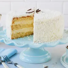 Eine köstliche Kokos Torte, bedeckt mit Kokosraspeln und dekoriert mit Kokoschips, wartet auf dem hellblauen Tortenständer.
