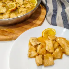 Petto di pollo al limone cremoso servito su un piatto bianco, con una fetta di limone e una padella piena dello stesso piatto sullo sfondo.