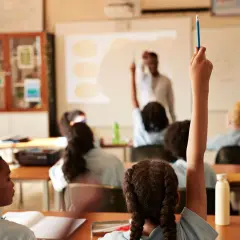 Estudiantes levantan la mano en clase. El profesor proyecta información, quizás sobre el calendario escolar 2026-27.