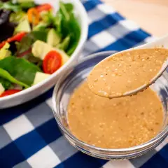 A spoon lifts creamy roasted sesame dressing from a bowl, ready to dress a fresh salad with avocado and tomatoes.