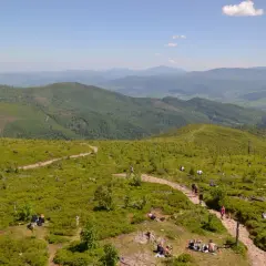 Widok z **przełęczy Kubalonka** na zielone, pofałdowane wzgórza Beskidów. Na ścieżkach i trawiastych zboczach odpoczywają turyści.