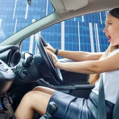 Mujer con expresión de susto al volante, parece que escucha un chirrido al girar el volante mientras conduce frente a un edificio moderno.