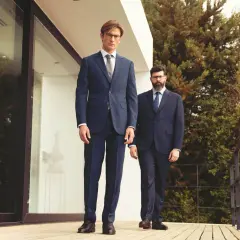 Deux hommes en costume sans chaussette, l'un grand et mince, l'autre plus trapu, posent sur une terrasse en bois devant une maison moderne.