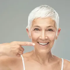 Donna sorridente indica il suo sorriso, un esempio di come rinforzare i denti a cinquant'anni con un sorriso smagliante.