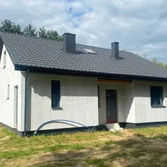 Nowy, biały dom szkieletowy z szarym dachem i dwoma kominami, otoczony zielenią.
