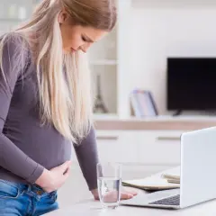 Kobieta w ciąży odczuwa skurcze przepowiadające, trzymając się za brzuch. Obok niej stoi szklanka wody i laptop.